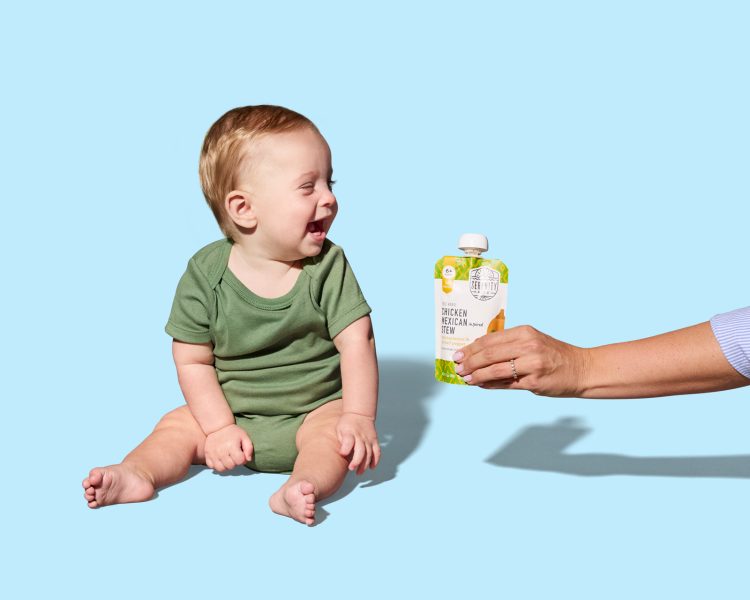 baby boy sitting on blue background with head turned smiling at hand with pouch