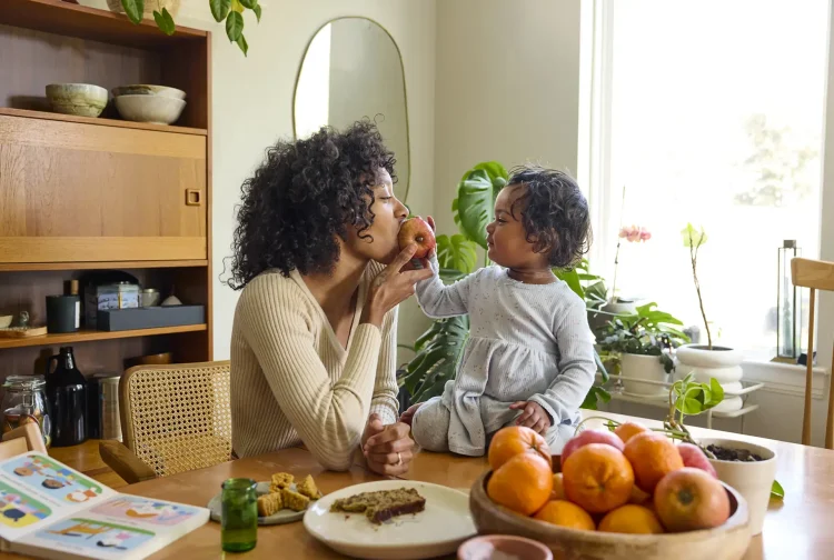 Mom and daughter share an apple at the kitchen table surrounded by a book, bread, and oranges