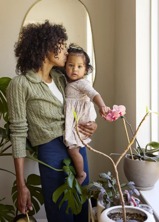 Mom holds daughter who's touching a pink orchid while watering the plants